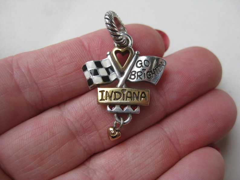 May include: A silver and gold charm with the word "INDIANA" on a gold rectangular plate. The charm features crossed racing flags, a heart-shaped cutout, and the words "GO BRIGHTON". A small gold heart dangles below.