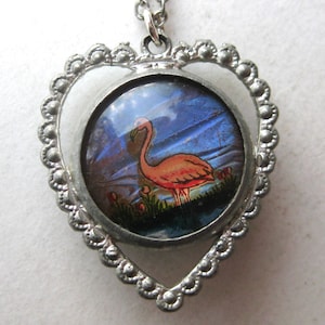 May include: A silver-toned heart-shaped pendant featuring a circular inset with a graphic illustration of a pink flamingo standing in water against a dark blue background. The metal frame has a scalloped edge and hangs from a silver-toned chain.