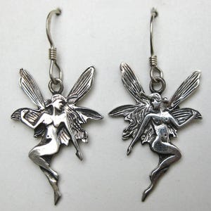 May include: A pair of silver-toned dangle earrings featuring a detailed fairy figure with outstretched wings. Each earring is suspended from a curved hook wire, showcasing a polished metallic finish with dark recessed areas to highlight the design.