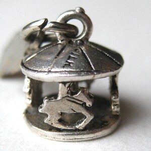 May include: A silver-toned metal charm shaped like a miniature carousel with a circular roof and a horse figure. The charm features a small jump ring at the top for attachment. The metal has a weathered, textured finish with visible dark patina.