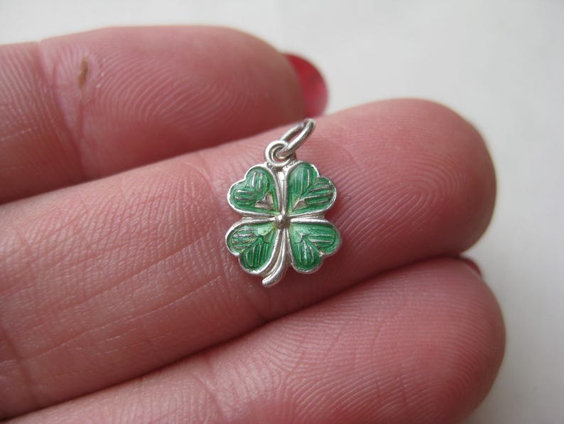 K&ouml;nnte beinhalten: Ein kleiner, silberfarbener Anh&auml;nger in Form eines vierbl&auml;ttrigen Kleeblatts. Die Bl&auml;tter des Kleeblatts sind mit gr&uuml;ner Emaille verziert, und der Anh&auml;nger hat eine kleine Schlaufe zum Befestigen an einer Halskette oder einem Armband. Ein Gl&uuml;ckssymbol.