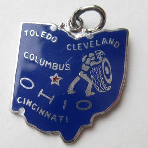 Könnte beinhalten: Ein silberfarbener Metallanhänger in Form des Bundesstaates Ohio mit blauer Emaille-Oberfläche. Er zeigt den Text Toledo, Cleveland, Columbus, Ohio und Cincinnati, einen kleinen roten Stern sowie die Grafik einer Person, die einen Reifen rollt.