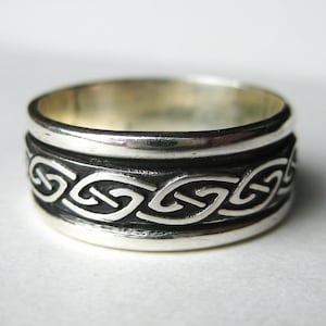May include: A silver ring with a Celtic knot design. The ring has a wide band with a raised border and a detailed pattern of interwoven knots. The ring is made of a shiny, reflective metal.
