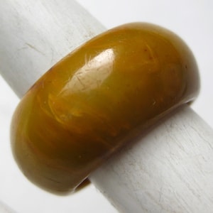 May include: A smooth, wide band ring in a solid mustard yellow color with a glossy, polished finish. The ring features subtle, swirling tonal variations throughout its surface, creating a marbled appearance across the rounded, thick band structure.