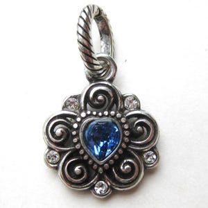 May include: A silver-toned metal pendant featuring a central heart-shaped blue crystal surrounded by ornate scrollwork and four small clear round crystals. The pendant includes a twisted rope-textured bail at the top for attachment to a chain.