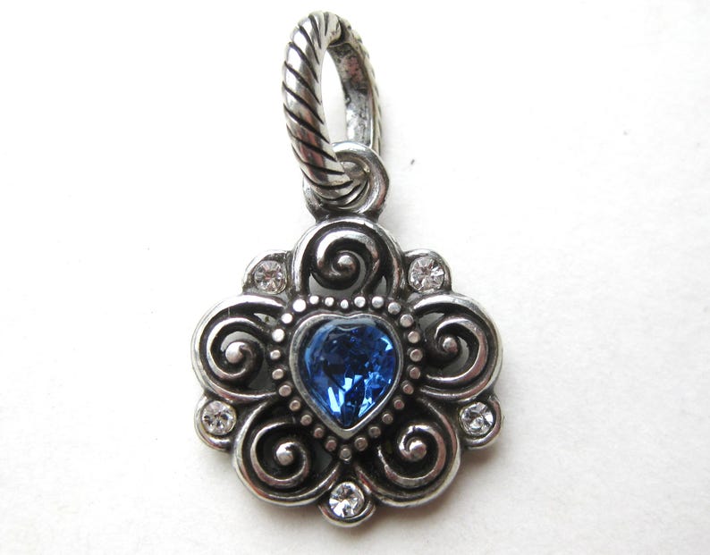 May include: A silver-toned metal pendant featuring a central heart-shaped blue crystal surrounded by ornate scrollwork and four small clear round crystals. The pendant includes a twisted rope-textured bail at the top for attachment to a chain.