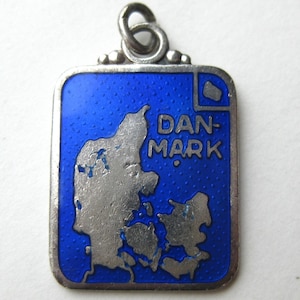 Könnte beinhalten: Rechteckiger Anhänger mit silberfarbenem Rand und blauem Emaille-Hintergrund. Der Anhänger zeigt eine Karte von Dänemark in Silber und das Wort "DAN-MARK" in weißen Buchstaben. Oben befindet sich eine kleine Schlaufe.