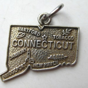May include: A silver-toned charm in the shape of the state of Connecticut. The charm features raised lettering of the state name and city names, including Hartford, Danbury, and New Haven. A small loop is attached for hanging.