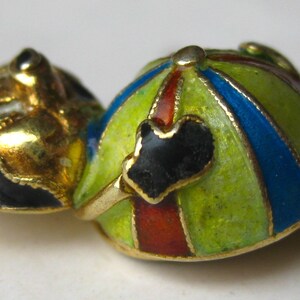 May include: A gold-toned metal charm featuring a stylized figure with a head and a rounded body. The body is decorated with enamel stripes in lime green, blue, and red. A small black heart-shaped detail is positioned on the front of the body.