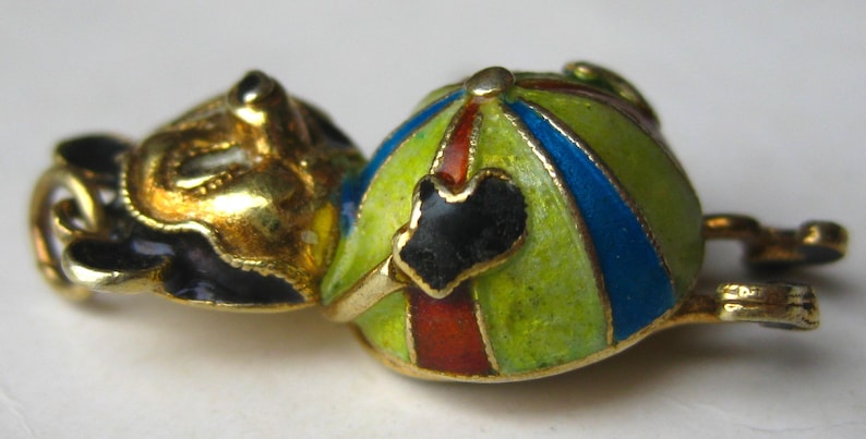 May include: A gold-toned metal charm featuring a stylized figure with a head and a rounded body. The body is decorated with enamel stripes in lime green, blue, and red. A small black heart-shaped detail is positioned on the front of the body.