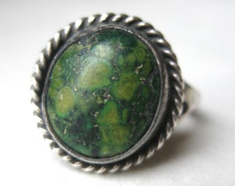 May include: A silver ring featuring a round, mottled green stone with dark inclusions. The stone is set within a polished silver bezel surrounded by a decorative twisted rope-style metal border, creating a textured frame around the center.