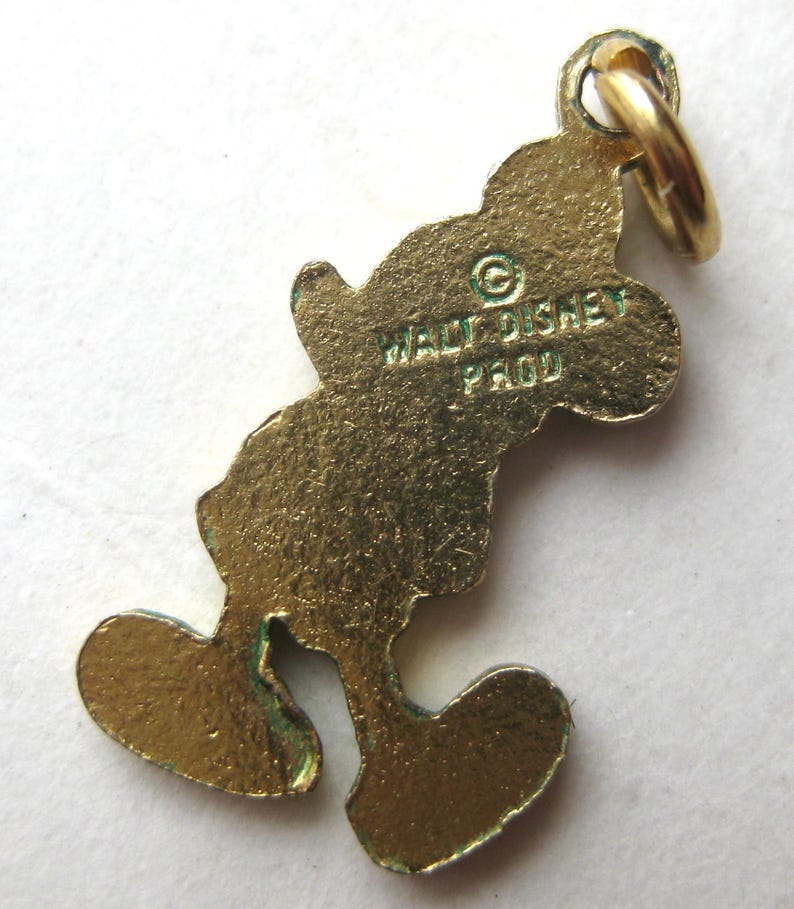 Puede incluir: Un charm de Mickey Mouse de color dorado con un lazo para sujetarlo a una pulsera o collar. El charm tiene el texto "WALT DISNEY PROD" en la parte posterior. El charm tiene la forma de Mickey Mouse.
