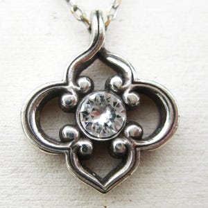 May include: A silver-toned pendant featuring a quatrefoil-shaped frame with oxidized detailing and a single, round, clear faceted gemstone set in the center. The pendant is attached to a thin silver-toned chain that extends upward from the top.