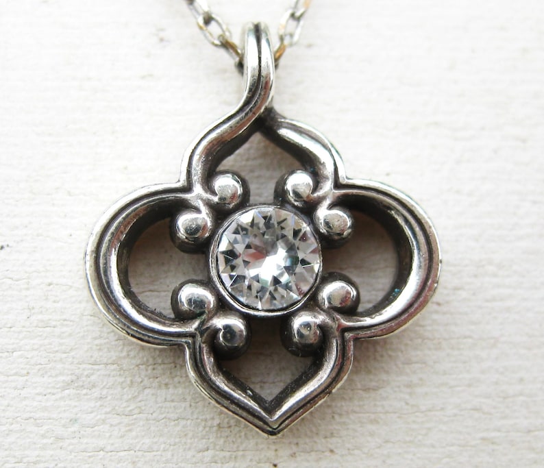 May include: A silver-toned pendant featuring a quatrefoil-shaped frame with oxidized detailing and a single, round, clear faceted gemstone set in the center. The pendant is attached to a thin silver-toned chain that extends upward from the top.