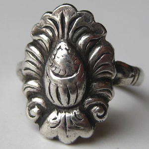 May include: A silver ring with an ornate, floral design. The ring features a central, textured element surrounded by stylized leaves and scrolls. The metal has a slightly aged appearance, enhancing the vintage aesthetic.