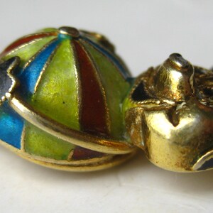May include: A gold-toned metal brooch shaped like a figure with a round body featuring segmented enamel panels in lime green, blue, and dark red. The figure has a gold head with black enamel accents on the ears and limbs, creating a stylized character.