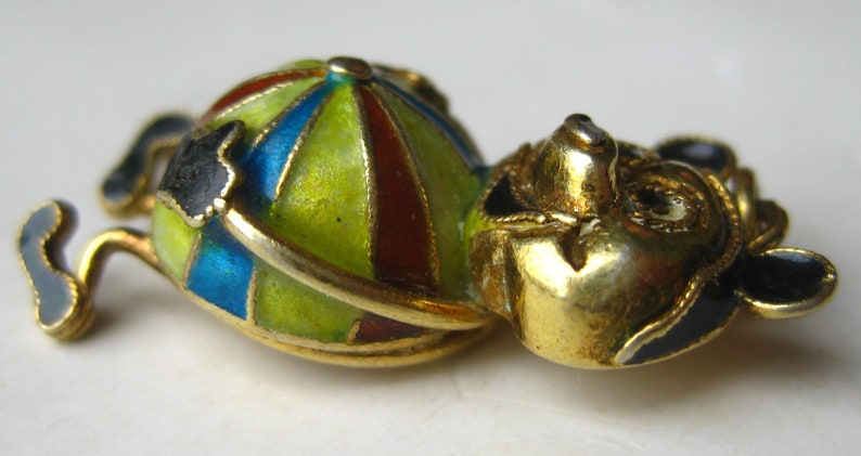 May include: A gold-toned metal brooch shaped like a figure with a round body featuring segmented enamel panels in lime green, blue, and dark red. The figure has a gold head with black enamel accents on the ears and limbs, creating a stylized character.