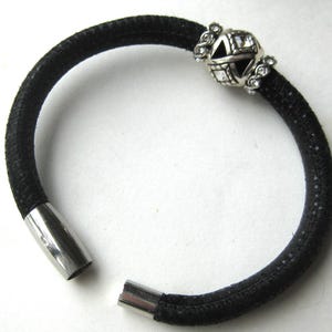 Puede incluir: Una pulsera negra texturizada que presenta una cuenta de metal plateado con cristales transparentes y recortes geom&eacute;tricos. La pulsera tiene un cierre magn&eacute;tico cil&iacute;ndrico de metal plateado liso en los extremos abiertos para un cierre seguro.