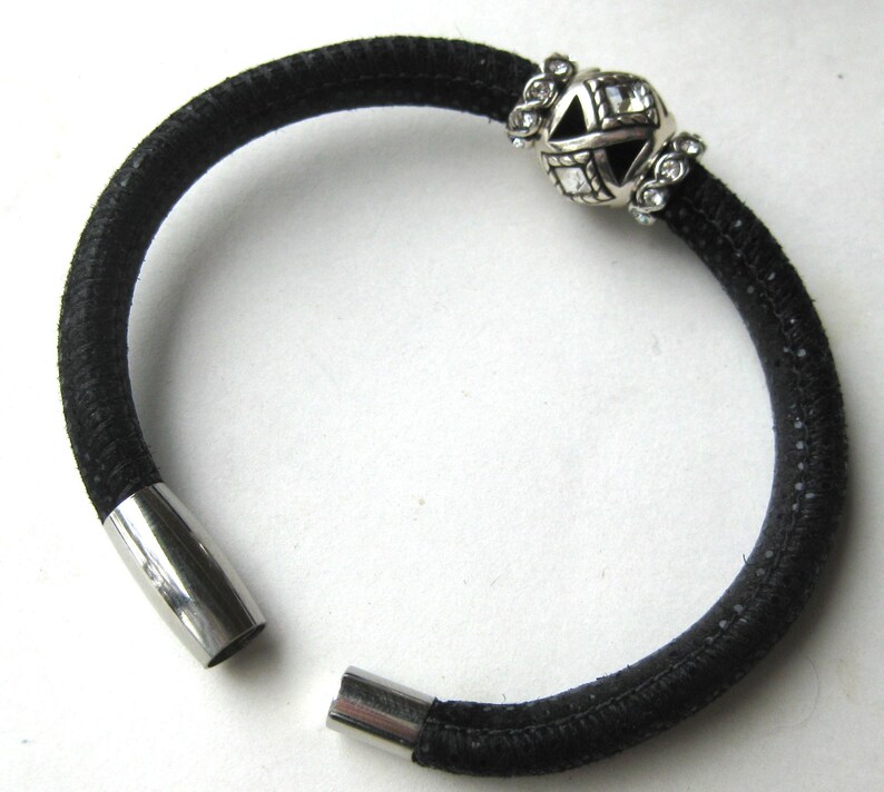 Puede incluir: Una pulsera negra texturizada que presenta una cuenta de metal plateado con cristales transparentes y recortes geom&eacute;tricos. La pulsera tiene un cierre magn&eacute;tico cil&iacute;ndrico de metal plateado liso en los extremos abiertos para un cierre seguro.