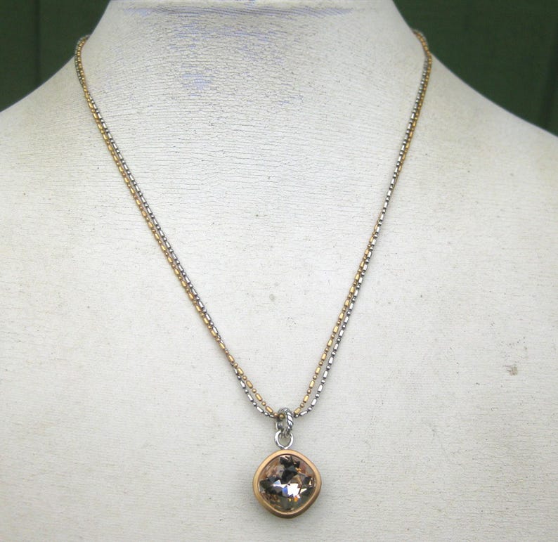 Puede incluir: Un collar con una cadena de dos tonos con cuentas alternas de color dorado y plateado. El colgante es un cristal redondo facetado de color champ&aacute;n engastado en un bisel de metal dorado, suspendido de una anilla de metal plateado.
