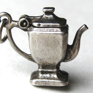 May include: A small, silver-toned metal charm shaped like a miniature teapot or samovar. The object features a square-based body, a curved handle with an attached jump ring, a short spout, and a lid with a small knob on top.