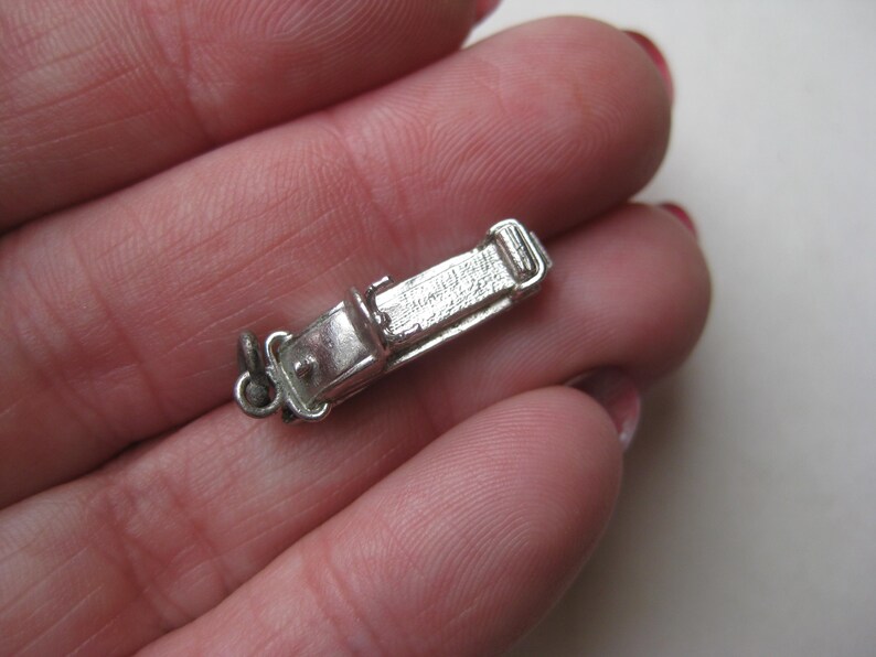 Może przedstawiać: Mały, srebrny charms w kształcie zabytkowego samochodu. Charms ma szczeg&oacute;łowe cechy, w tym dach, okna i koła. Jest przymocowany do małej pętli, co sugeruje, że może być używany jako wisiorek lub zawieszka do bransoletki.