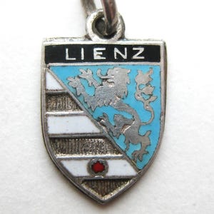 May include: A silver-toned pendant with a shield design. The shield features the word "LIENZ" in black, a light blue background with a silver lion, and white and silver stripes. A red dot is at the bottom.