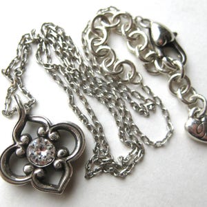 May include: A silver-toned necklace featuring a quatrefoil-shaped pendant with a central clear round crystal. The chain includes a lobster claw clasp and a small heart-shaped charm at the end, with a series of circular links for length adjustment.