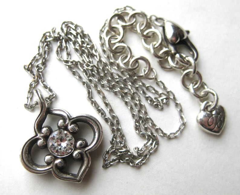 May include: A silver-toned necklace featuring a quatrefoil-shaped pendant with a central clear round crystal. The chain includes a lobster claw clasp and a small heart-shaped charm at the end, with a series of circular links for length adjustment.