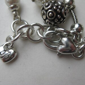 Puede incluir: Primer plano de un cierre y cadena de collar plateados. El cierre tiene forma de coraz&oacute;n, y un peque&ntilde;o dije de coraz&oacute;n con un dise&ntilde;o grabado cuelga de la cadena. El collar presenta cuentas plateadas y una bola decorativa.