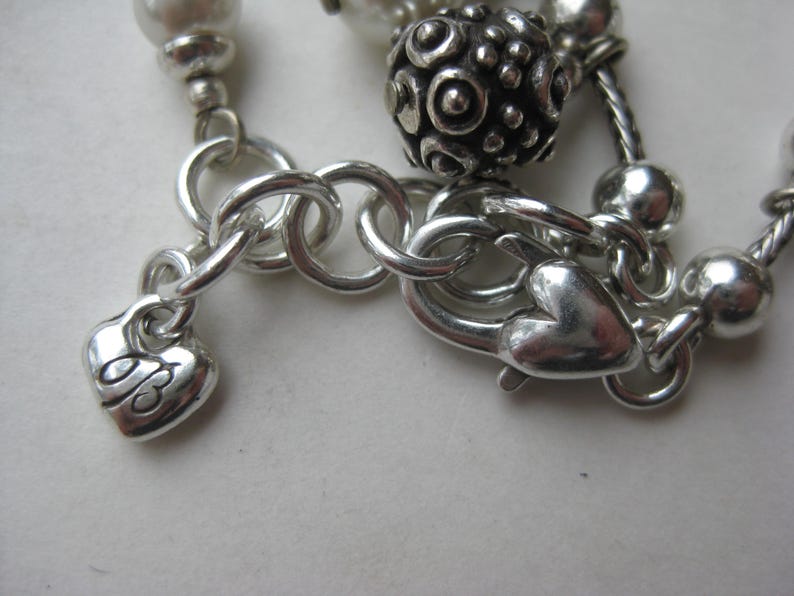 Puede incluir: Primer plano de un cierre y cadena de collar plateados. El cierre tiene forma de coraz&oacute;n, y un peque&ntilde;o dije de coraz&oacute;n con un dise&ntilde;o grabado cuelga de la cadena. El collar presenta cuentas plateadas y una bola decorativa.