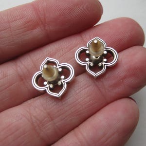 May include: A pair of silver-toned stud earrings featuring a four-lobed, openwork quatrefoil design. Each earring has a central post with a translucent, flexible plastic backing attached, showcasing a metallic finish with dark recessed detailing.