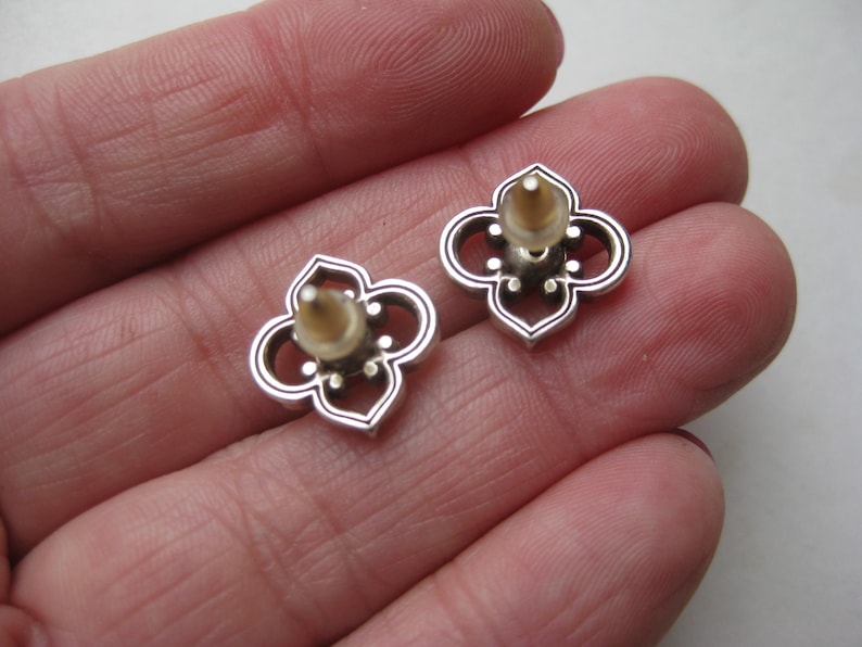 May include: A pair of silver-toned stud earrings featuring a four-lobed, openwork quatrefoil design. Each earring has a central post with a translucent, flexible plastic backing attached, showcasing a metallic finish with dark recessed detailing.