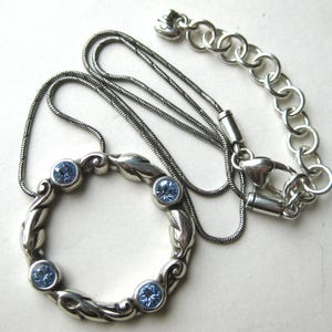 May include: Silver-toned necklace featuring a circular pendant with four light blue gemstones. The pendant has a decorative, swirling design. The necklace chain is a darker silver color and includes an adjustable chain with a heart-shaped clasp.