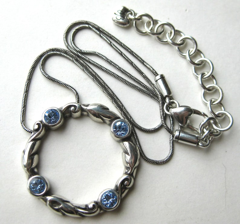 May include: Silver-toned necklace featuring a circular pendant with four light blue gemstones. The pendant has a decorative, swirling design. The necklace chain is a darker silver color and includes an adjustable chain with a heart-shaped clasp.