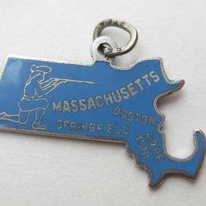 May include: A silver-toned Massachusetts charm with a blue enamel finish. The charm is shaped like the state and features the word "Massachusetts" and the cities of Boston, Springfield, and Cape Cod. A silver-toned figure of a person with a rifle is also depicted.