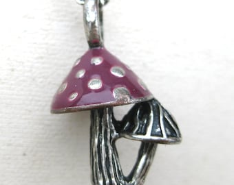 May include: A silver-toned metal pendant featuring two mushrooms. The larger mushroom has a deep purple enamel cap with silver polka dots, while the smaller mushroom is solid silver-toned metal with vertical ridges on the cap and stem.