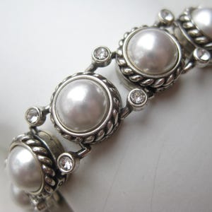 Puede incluir: Una pulsera de tono plateado que presenta una serie de perlas blancas redondas engastadas en bordes de metal trenzado. Cada engaste de perla est&aacute; conectado por peque&ntilde;os cristales transparentes de corte redondo, creando un patr&oacute;n de eslabones.