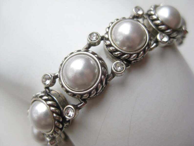 Puede incluir: Una pulsera de tono plateado que presenta una serie de perlas blancas redondas engastadas en bordes de metal trenzado. Cada engaste de perla est&aacute; conectado por peque&ntilde;os cristales transparentes de corte redondo, creando un patr&oacute;n de eslabones.