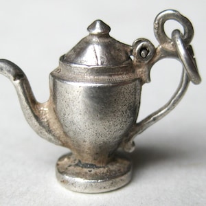 May include: A miniature silver-toned metal teapot charm featuring a curved spout, a hinged lid with a pointed top, and a handle attached to a circular jump ring. The surface has a weathered, metallic finish with visible signs of age and texture.