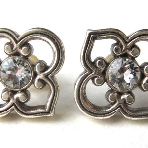May include: A pair of silver-toned stud earrings featuring a four-petaled openwork design with a circular clear crystal set in the center of each piece. The metal has a slightly darkened finish in the recessed areas to highlight the intricate shape.