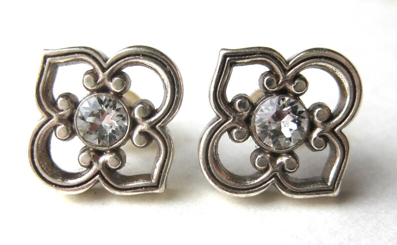 May include: A pair of silver-toned stud earrings featuring a four-petaled openwork design with a circular clear crystal set in the center of each piece. The metal has a slightly darkened finish in the recessed areas to highlight the intricate shape.