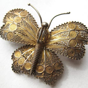 May include: A vintage butterfly brooch with intricate filigree detailing. The wings feature a gold-tone finish with silver-tone accents, and the body has a textured appearance. The brooch has delicate antennae and a pin closure on the back.