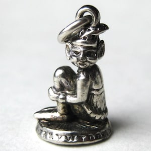 May include: A small silver charm depicting a seated elf-like figure with pointed ears, wearing a hat and a tunic. The figure is positioned on a circular base with a textured edge and includes a jump ring at the top for attachment to a bracelet or chain.