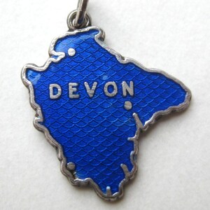 May include: A silver-toned pendant in the shape of the county of Devon, England. The pendant has a bright blue enamel finish with a scale-like texture. The word "DEVON" is displayed in silver letters across the center.