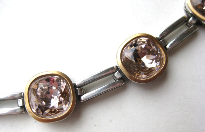 May include: A bracelet featuring a series of square-shaped, light champagne-colored faceted crystals. Each crystal is set within a smooth, matte gold-toned bezel, connected by polished silver-toned rectangular metal links in a chain design.