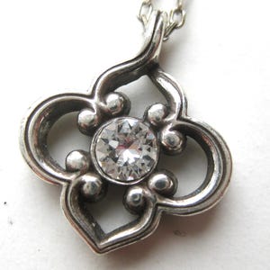 May include: A silver-toned pendant featuring a quatrefoil-shaped frame with rounded edges and a central, circular, clear faceted gemstone. The pendant is suspended from a delicate silver-toned chain, showcasing a polished metallic finish.