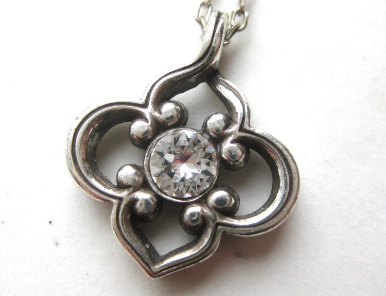 May include: A silver-toned pendant featuring a quatrefoil-shaped frame with rounded edges and a central, circular, clear faceted gemstone. The pendant is suspended from a delicate silver-toned chain, showcasing a polished metallic finish.
