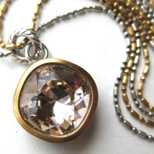 Puede incluir: Un collar con un gran colgante de cristal de color champ&aacute;n, de corte cuadrado, engastado en un bisel de tono dorado mate. El colgante cuelga de una cadena de m&uacute;ltiples hilos compuesta por cuentas y eslabones met&aacute;licos plateados y dorados.