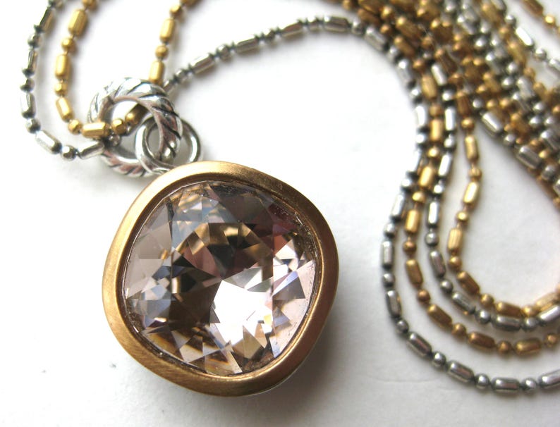 Puede incluir: Un collar con un gran colgante de cristal de color champ&aacute;n, de corte cuadrado, engastado en un bisel de tono dorado mate. El colgante cuelga de una cadena de m&uacute;ltiples hilos compuesta por cuentas y eslabones met&aacute;licos plateados y dorados.